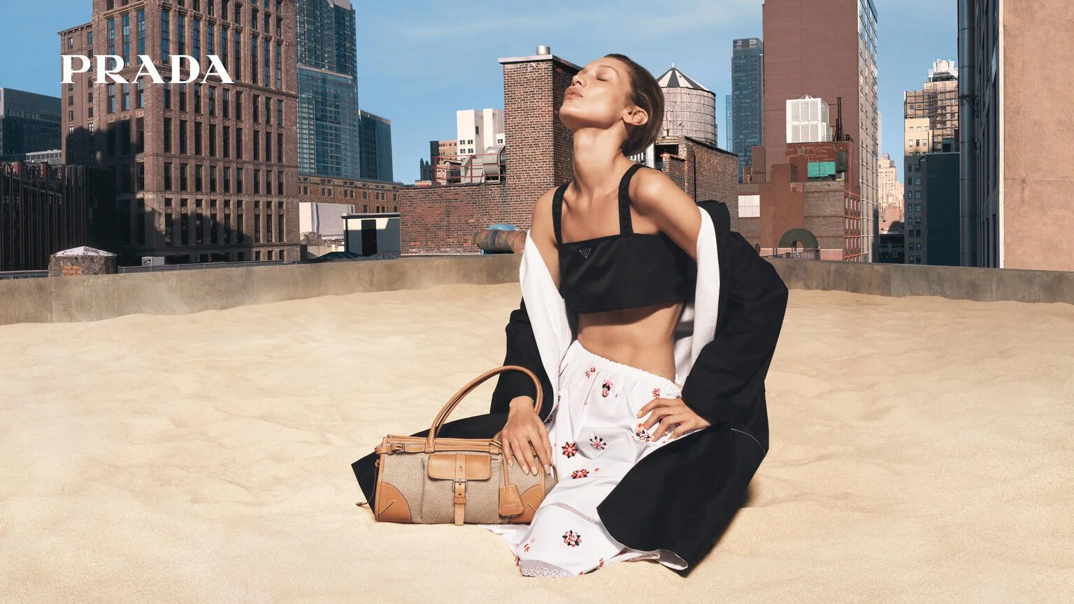 A woman with closed eyes and tilted head sits on a sandy rooftop in front of a city skyline with tall buildings, holding a beige handbag, wearing black and white clothes, and a black crop top.