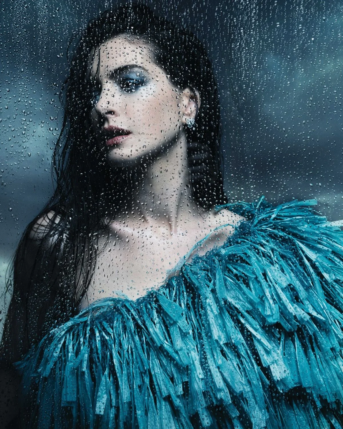A woman with dark wet hair and light makeup looks through a rain-covered glass with a cloudy, stormy sky in the background, wearing a textured blue dress.