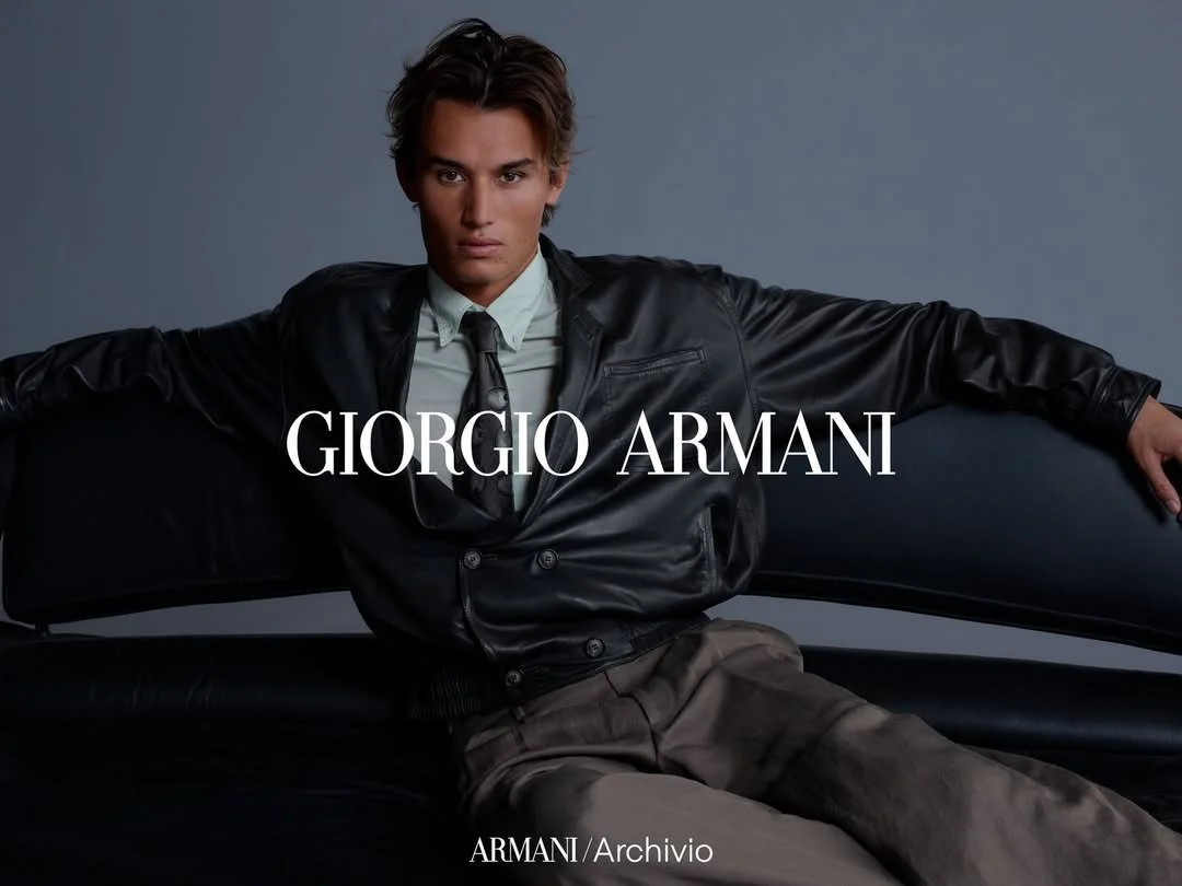 A young man in a black leather jacket, white shirt, and tie sitting on a black leather couch against a plain gray background with 'Giorgio Armani' branding overlayed in white text.