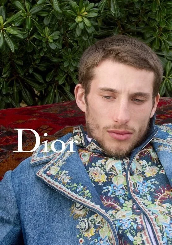 A young man with light brown hair and a beard, dressed in a floral embroidered denim jacket, sitting outdoors with green bushes in the background, and the word 'Dior' overlaid on the image.