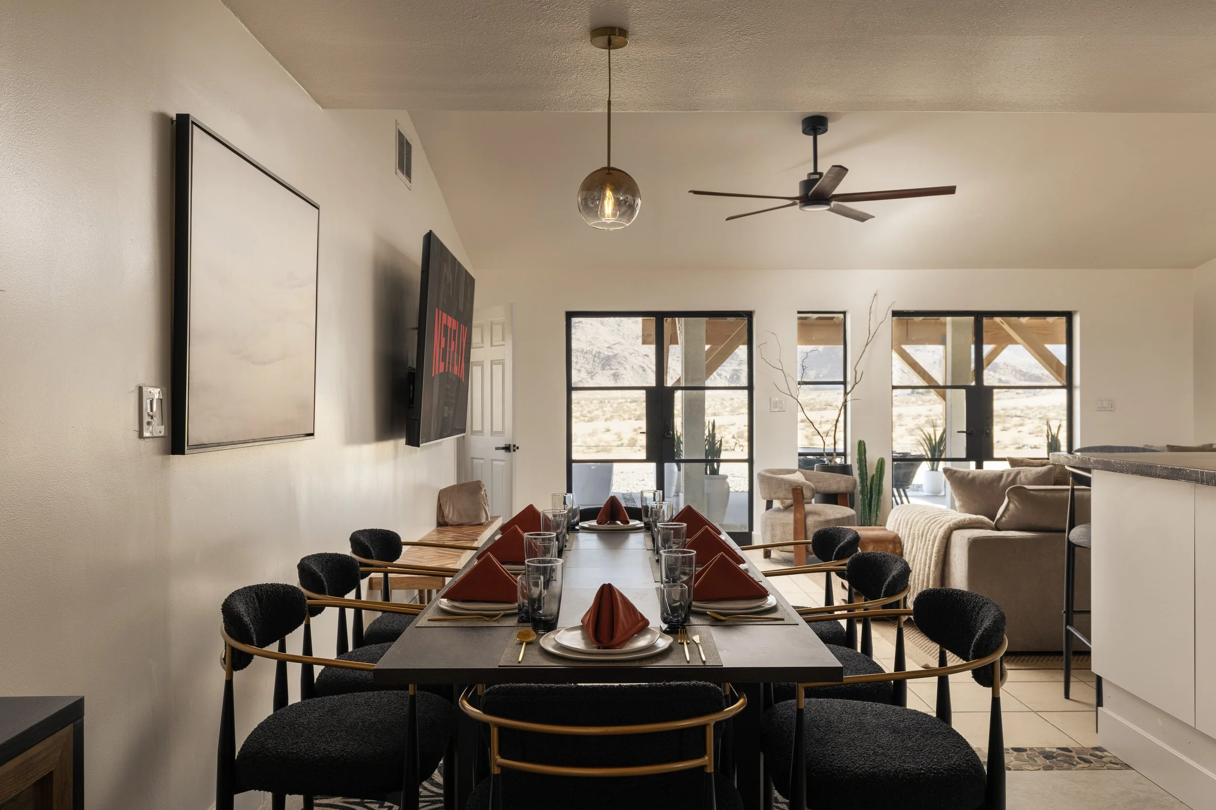 Interior of a modern dining and living area with a dining table set with glasses, plates, and napkins, letters, sofas, large windows, and ceiling fan.