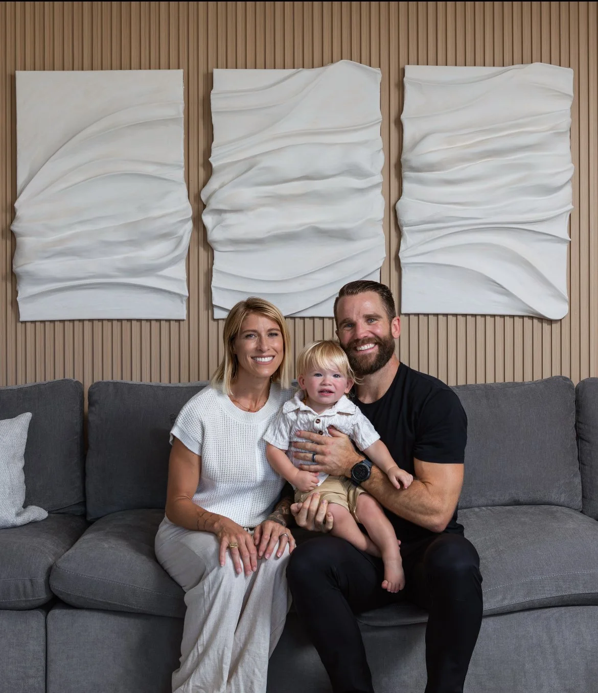 A family of three sitting on a gray couch in a living room, smiling at the camera. Behind them are three white abstract wall art pieces against a wooden panel wall. The woman has blonde hair and is wearing a white top, the man has a beard and is wearing a black t-shirt, and the child has blonde hair and is wearing a striped shirt and shorts.