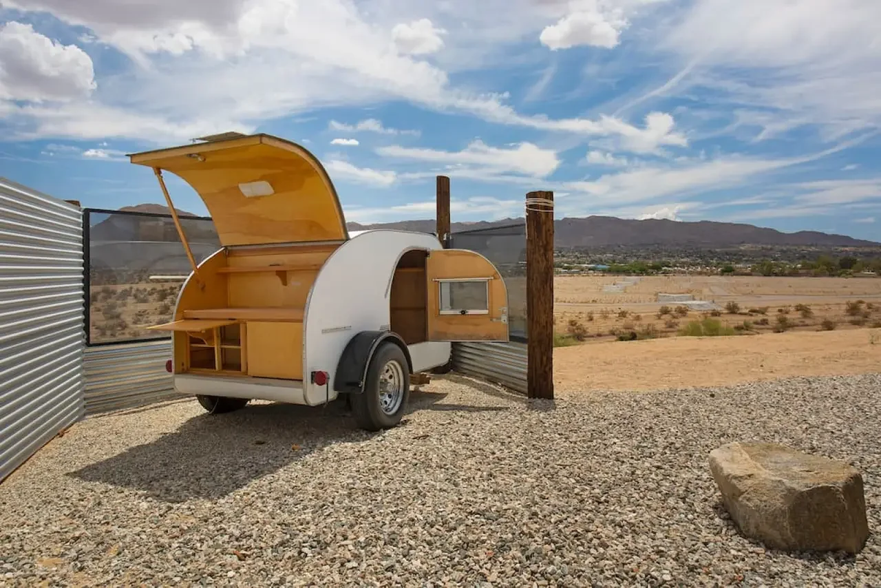 Small white trailer with a curved wooden interior and open top, parked on gravel in a desert landscape with mountains and blue sky in the background.