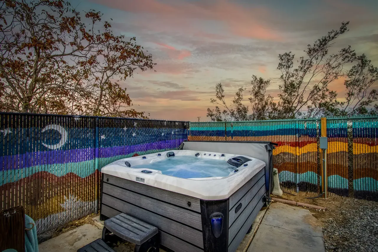 A hot tub filled with water and jets inside a fenced outdoor area at sunset, with trees and a colorful mural on the fence depicting mountains and sky.