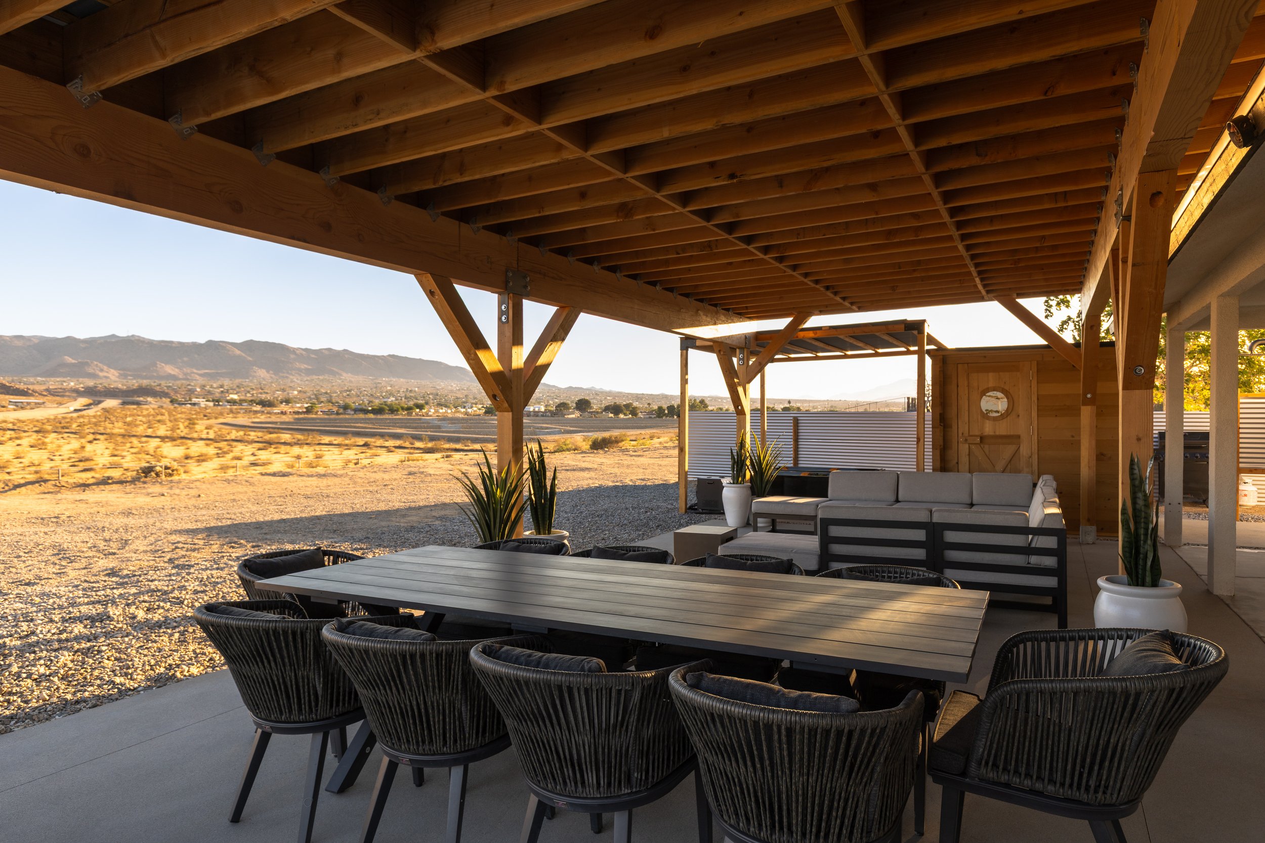 Outdoor patio area with a long dining table and black wicker chairs. Overhead, a wooden ceiling with cross beams is visible. In the background, there are trees and mountains under a clear sky.
