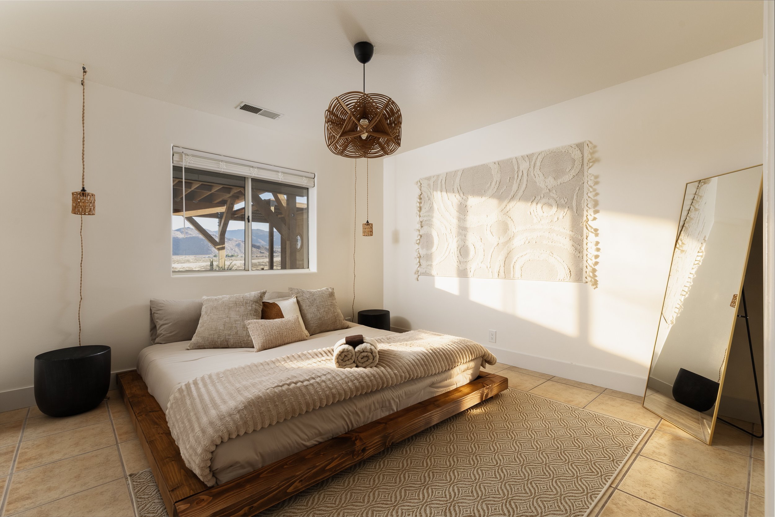 Sunlit bedroom with a low platform bed topped with beige and brown pillows and rolled towels, a large textured wall hanging, a tilted mirror, and a window revealing a desert landscape with mountains in the background.