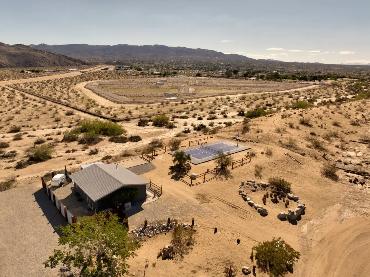 A desert landscape with sparse vegetation, a small building, a tennis court, a fire pit circle made of rocks, and surrounding hills with mountains in the background.