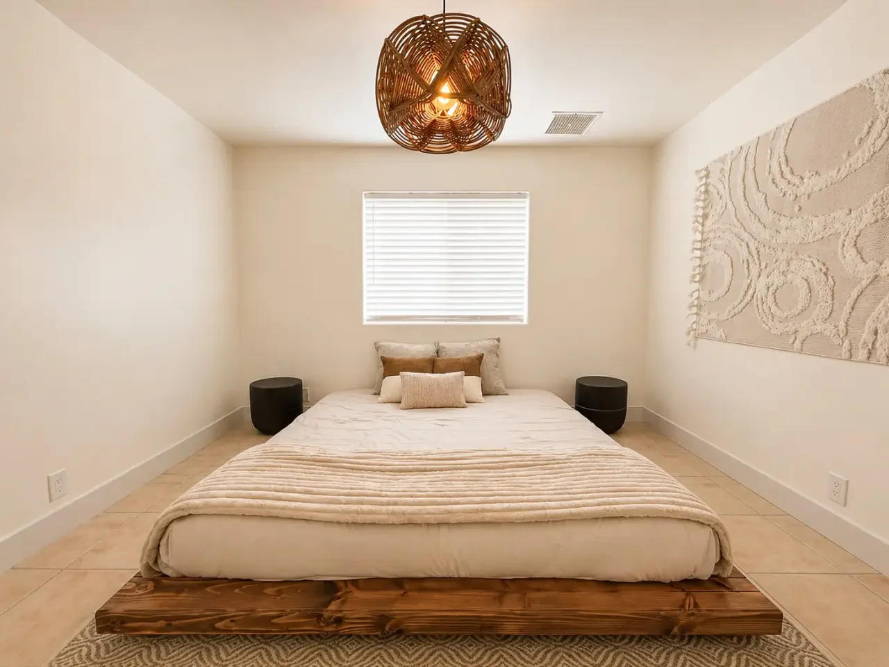 Minimalist bedroom with a low bed on a wooden platform, beige bedding, decorative pillows, black side tables, a window with closed blinds, a textured wall hanging, and a woven ceiling light fixture.