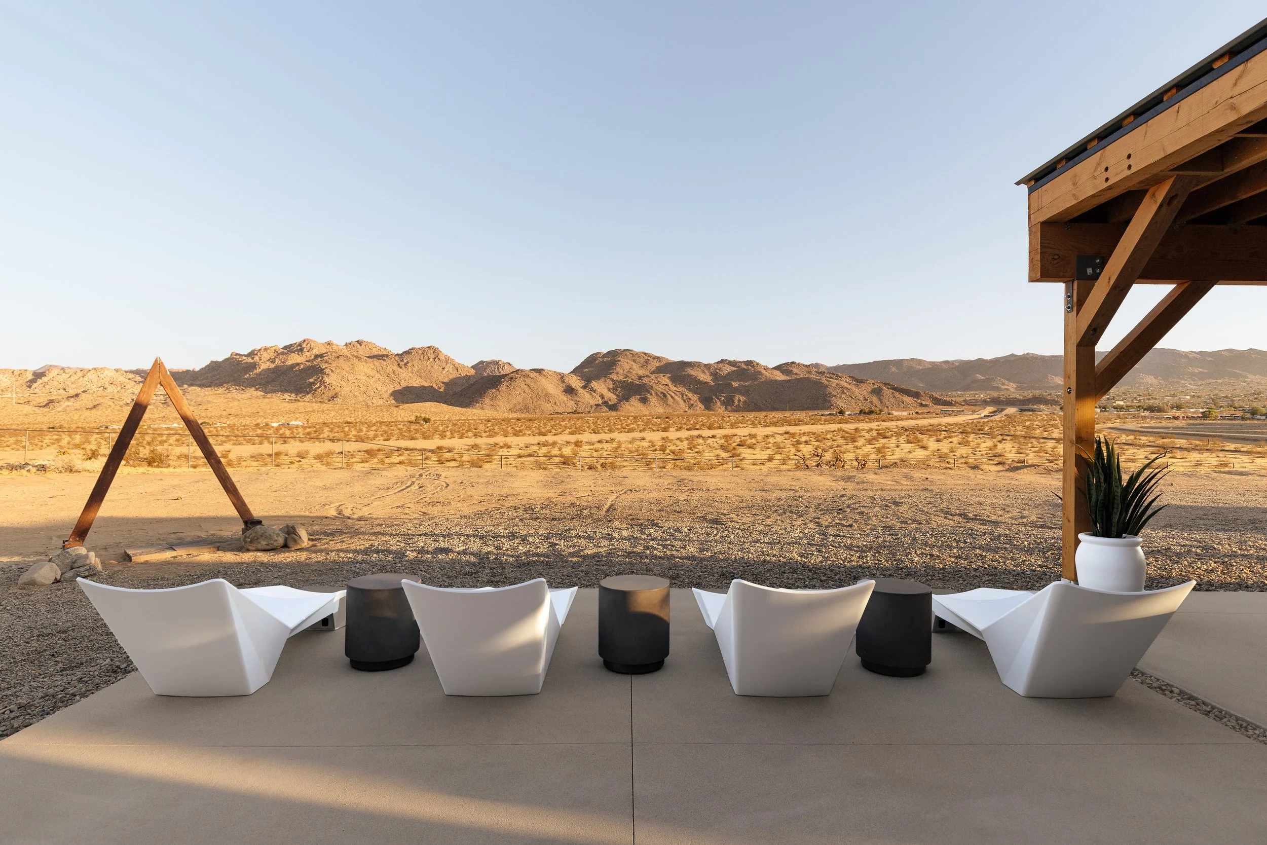 Outdoor seating area with modern white chairs and black stools on a concrete patio, overlooking a desert landscape with mountains in the background, under a clear sky.