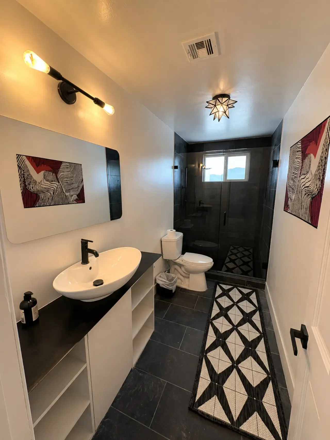 Modern bathroom with black and white decor, including a sink, mirror, toilet, walk-in shower with black tile, and decorative rugs.