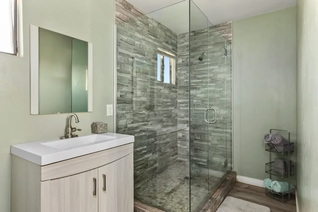Bathroom with a glass shower enclosure, a light-colored vanity with a sink and mirror, small window, and a black metal towel rack with rolled towels.