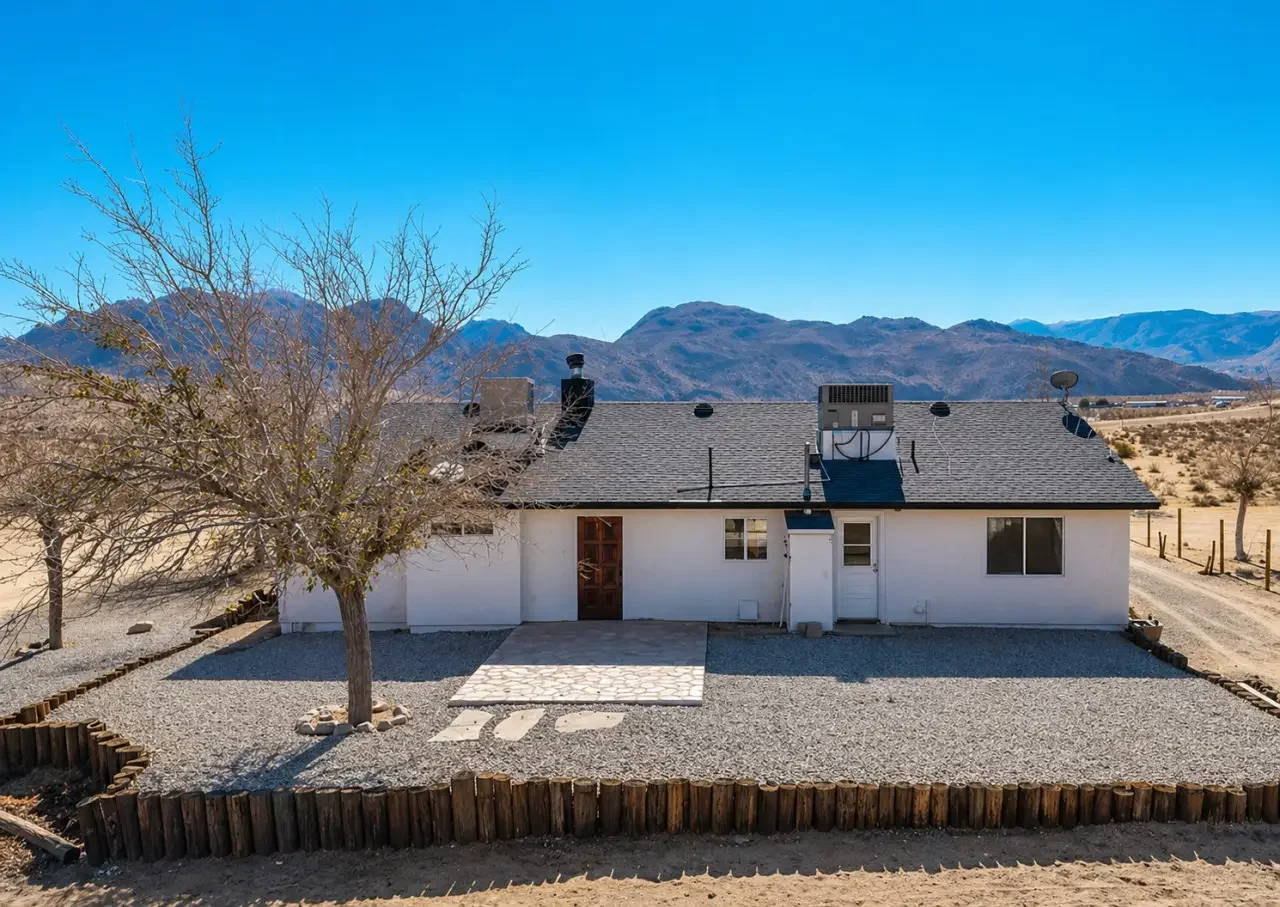 A white house with a gray shingle roof, surrounded by a gravel yard enclosed with wooden border, in a dry landscape with mountains in the background and a clear blue sky.