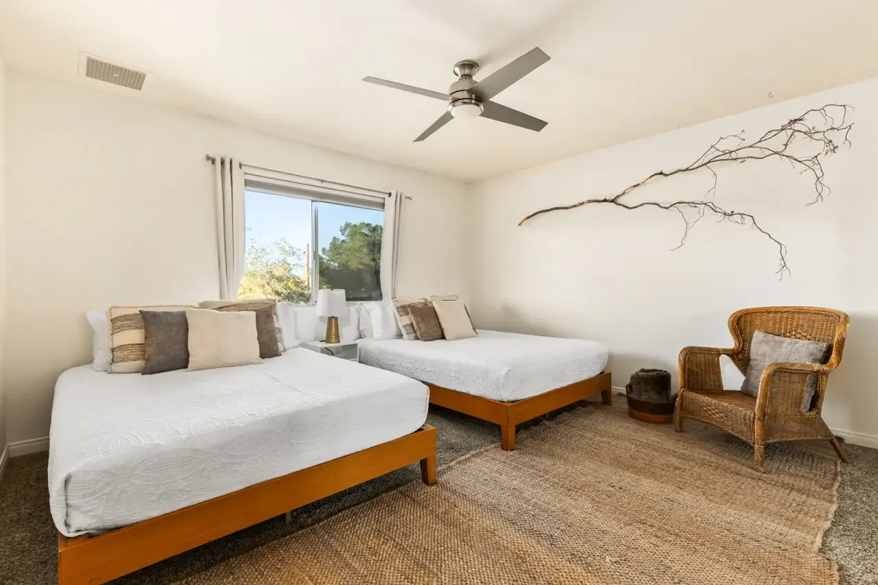 A bedroom with two beds, a wicker chair, a small table, and a window showing green trees outside. The room has a ceiling fan and a branch decor on the wall.