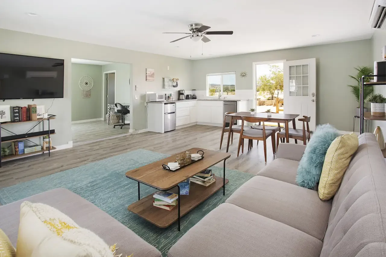 Open-concept living room and kitchen with light green walls, wood flooring, a ceiling fan, a large flat-screen TV, a gray sofa with decorative pillows, a coffee table with books and decor, a dining table with chairs, and a door leading outside.