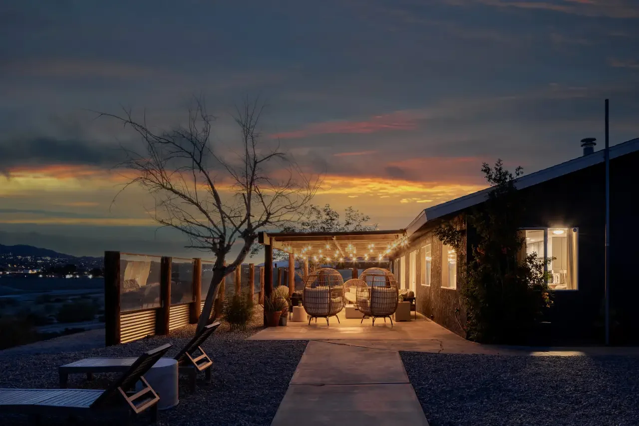A cozy outdoor patio with string lights, wicker chairs, and a view of a sunset over water in the evening.