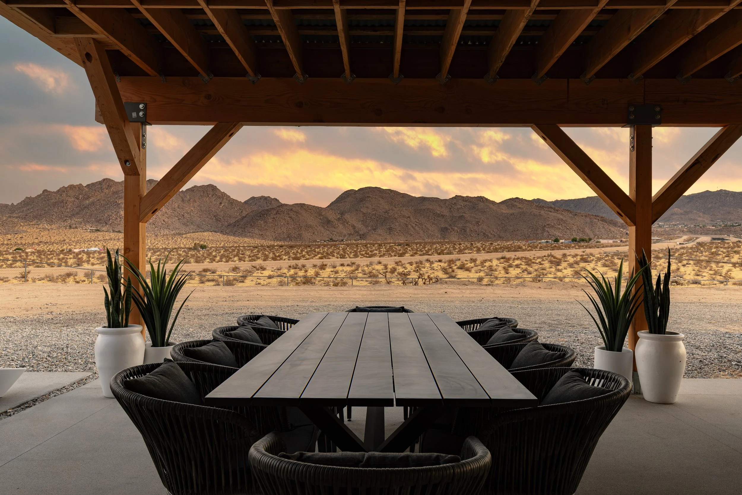 Outdoor patio area with a long table and black chairs, potted plants on each side, wooden beam structure overhead, desert landscape with mountains in the background at sunset.