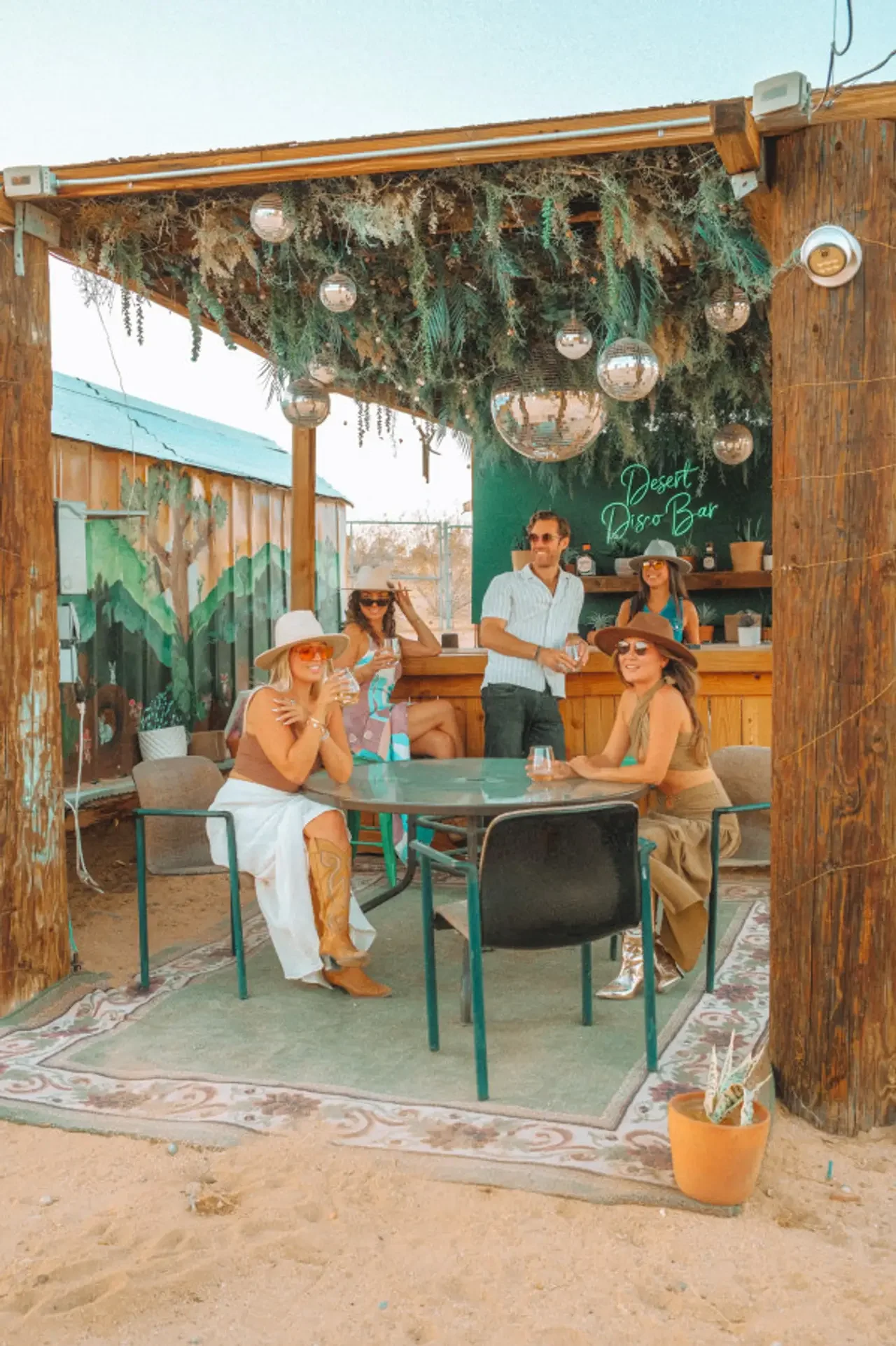 Group of five people enjoying drinks at an outdoor desert-themed bar with a green sign that reads 'Desert Disco Bar', wooden framing, greenery, and hanging silver spheres.