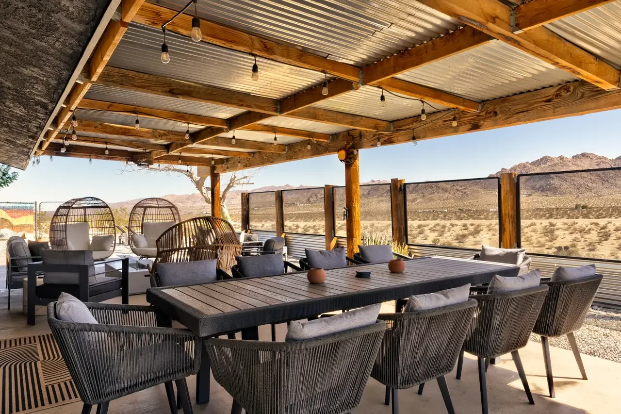 Outdoor patio with wooden table and chairs, hanging string lights, and desert landscape with mountains in the background.