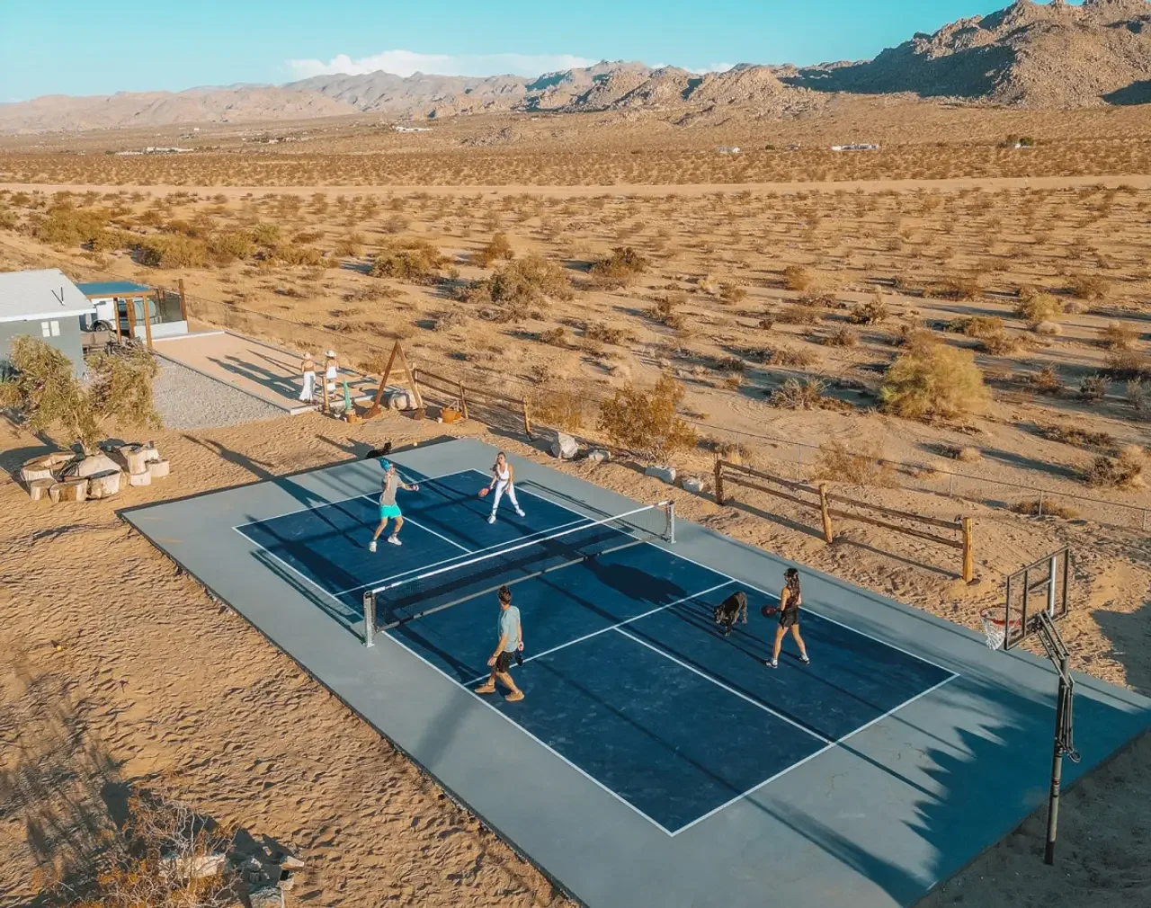 People playing tennis on an outdoor court in a desert landscape with mountains in the background.