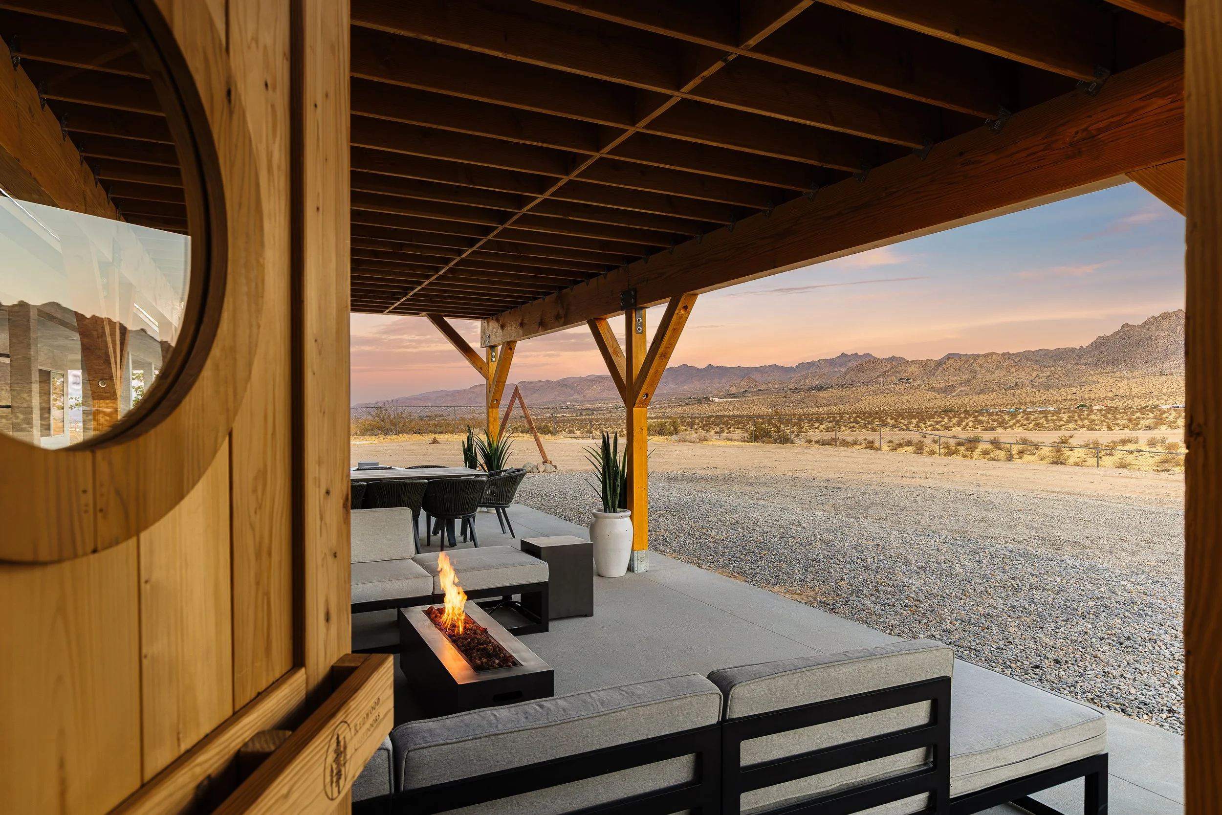 Covered outdoor patio area with modern seating and a linear fire feature, desert landscape and mountains in the distance at sunset.