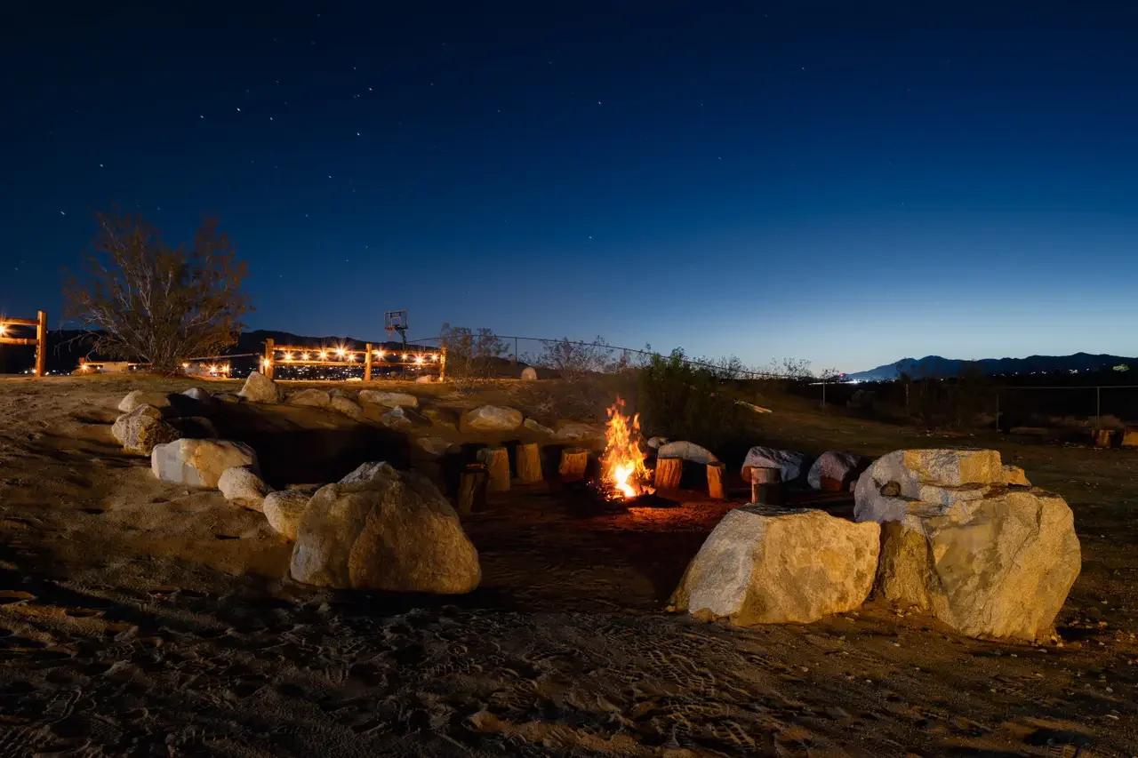 Nighttime outdoor scene with a small campfire surrounded by large rocks, in a desert-like setting with a fence, a tree, distant mountains, and a clear sky with stars.