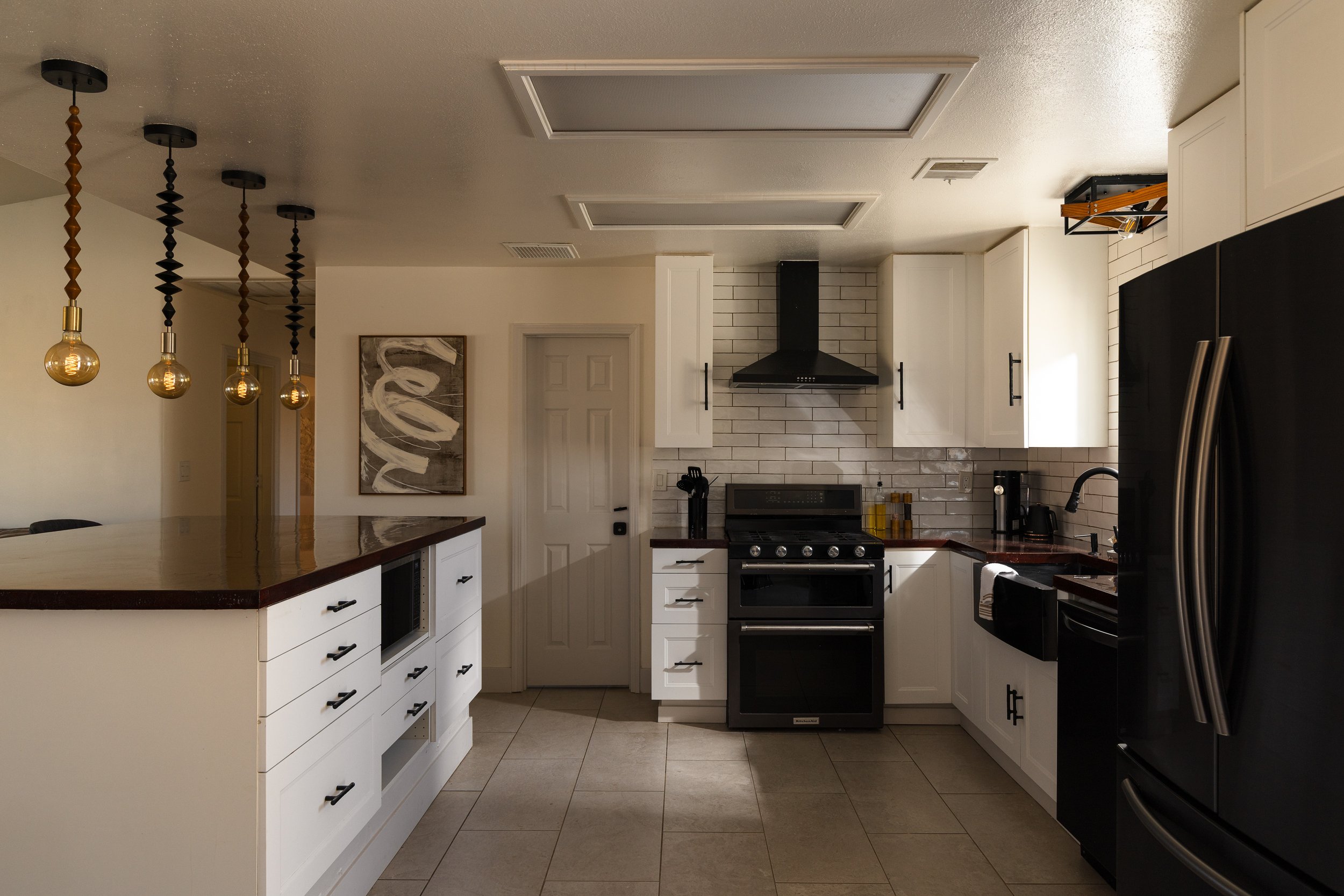Modern kitchen with white cabinets, black appliances, pendant lights, and artwork on the wall.