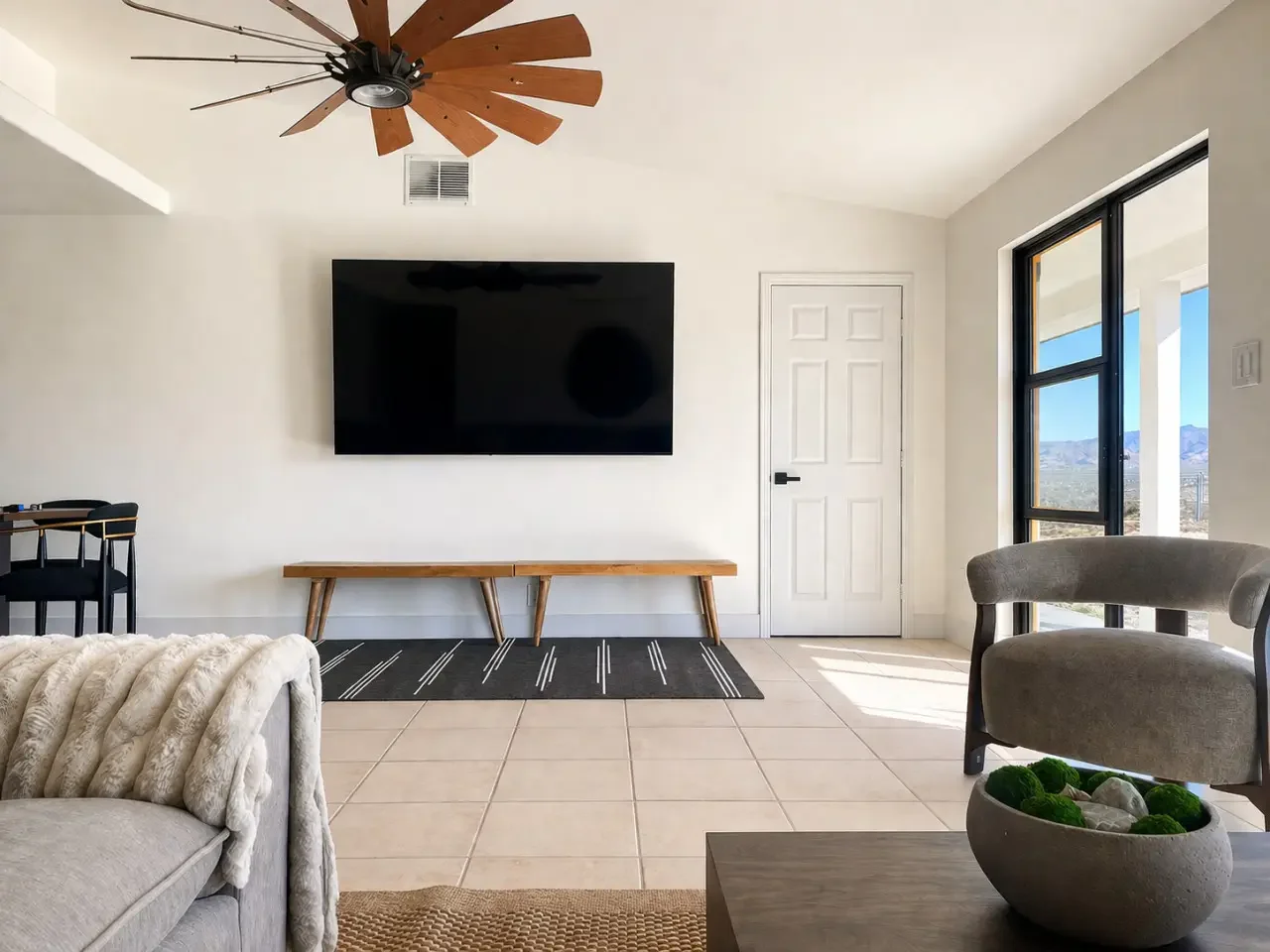 Living room with a wall-mounted flat-screen TV, a wooden bench underneath, a light-colored sofa with a plush throw blanket, a modern armchair, a side table with a decorative bowl, large sliding glass doors showing a desert landscape, and a ceiling fa