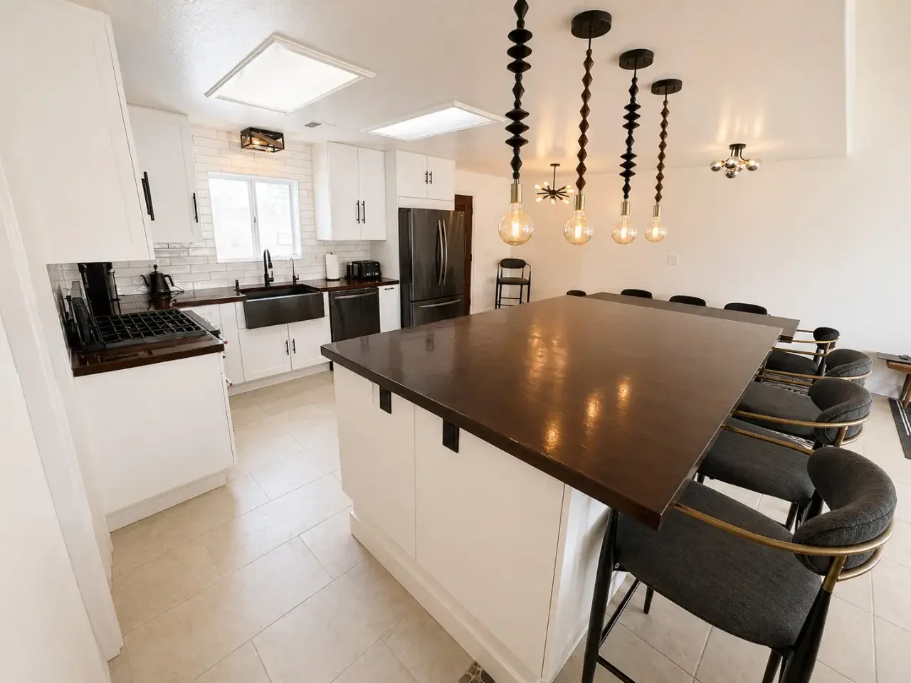 Modern kitchen with white cabinets, black hardware, a large dark wood island, black barstools, a stainless steel refrigerator, and pendant lights with exposed bulbs hanging above the island.