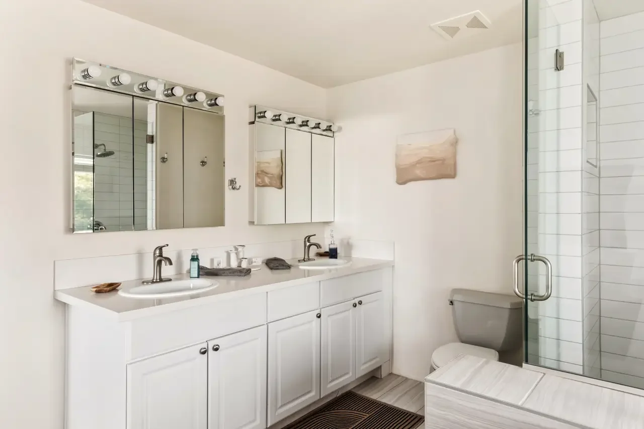 Modern bathroom featuring a double vanity with white cabinets and two sinks, a large framed mirror with light bulbs, a glass-enclosed shower, and a toilet, with minimal decorative accents.