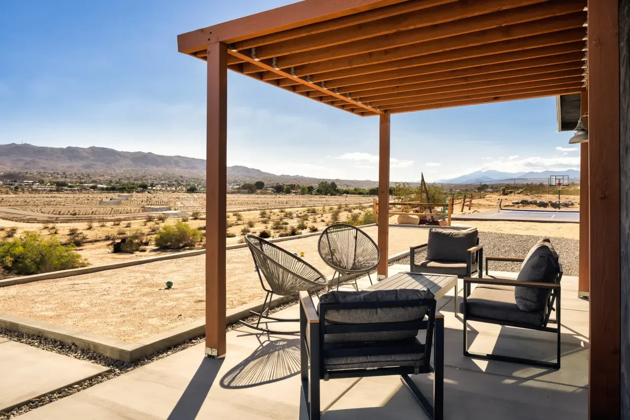 Patio with outdoor furniture under a wooden pergola, overlooking a desert landscape with mountains in the distance and a basketball hoop in the background.