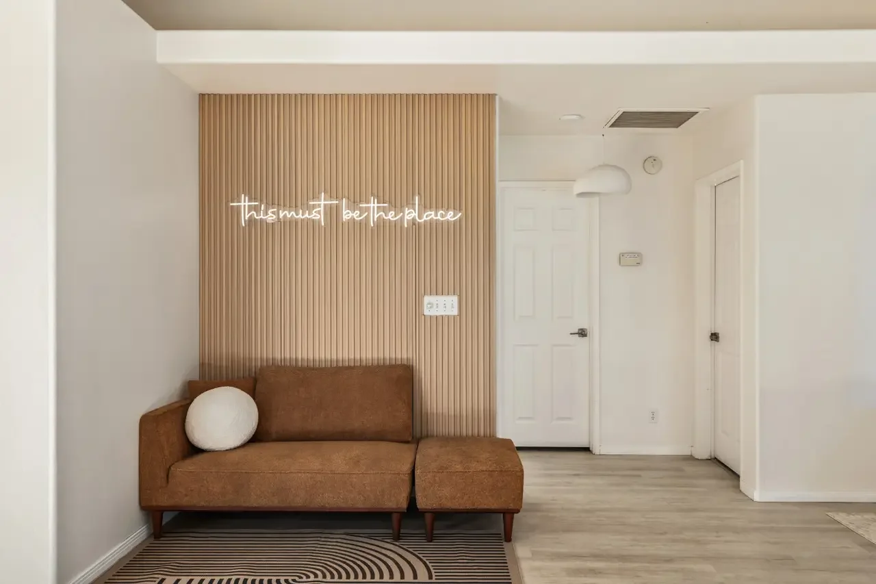 Living room area with a brown sofa, a round white pillow, a wooden panel wall with neon sign saying 'this must be the place,' a white door, and a beige rug with black lines.