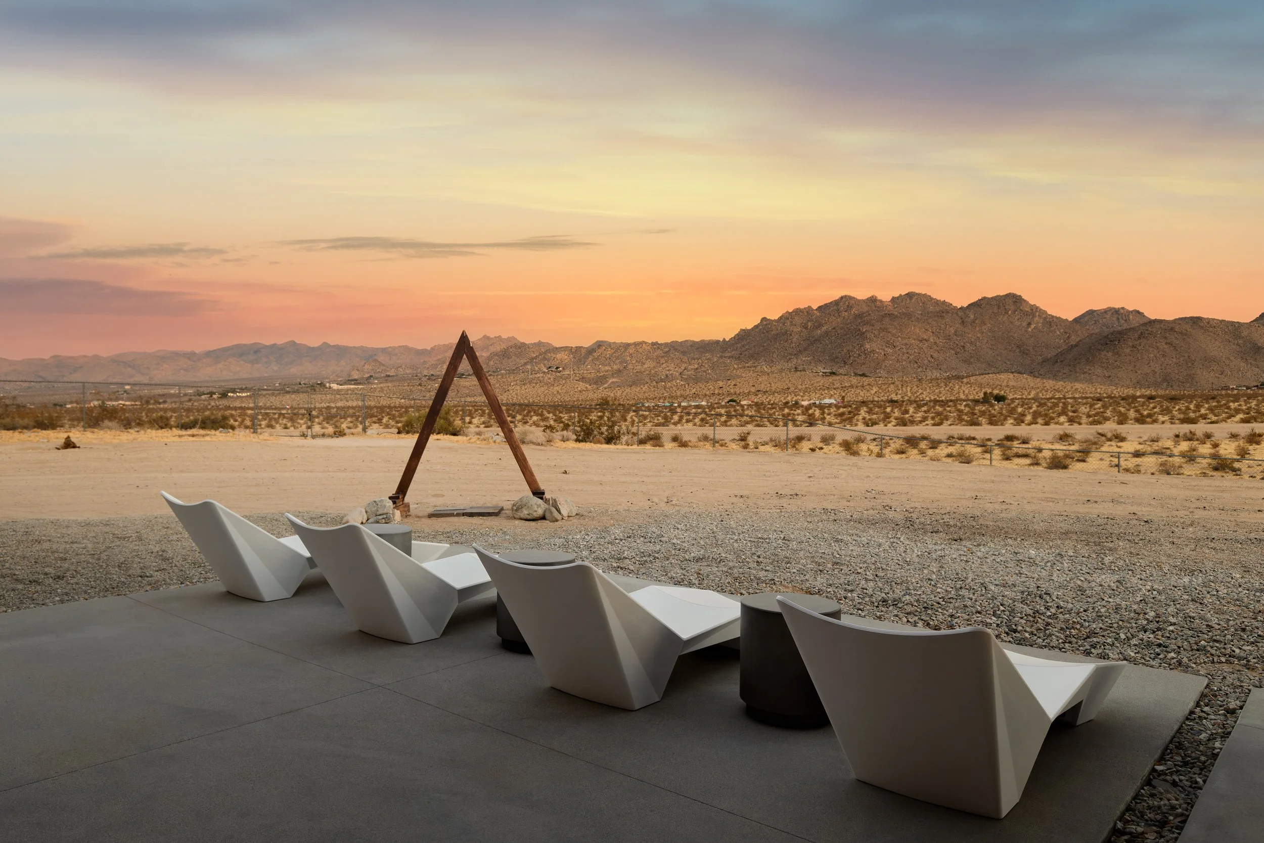 A desert landscape during sunset with mountains in the distance, a triangle-shaped wooden structure, and modern white lounge chairs on a patio.