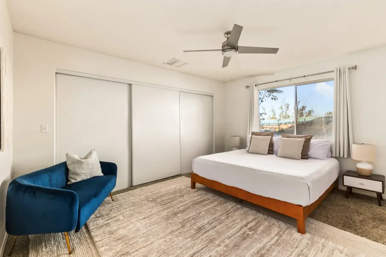 A bedroom with a bed, three pillows, a side table with a lamp, a closet with sliding doors, a window with white curtains, a ceiling fan, a blue velvet sofa with a throw pillow, and a beige area rug.