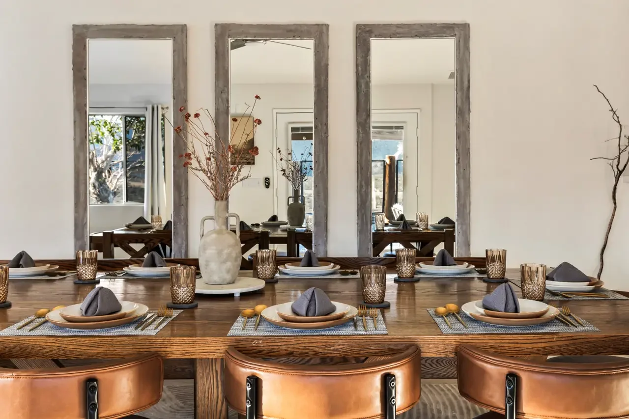 A dining table set with plates, napkins, glasses, and cutlery in a room with three large mirrors on the wall, a vase with dried flowers in the center, and windows showing trees outside.