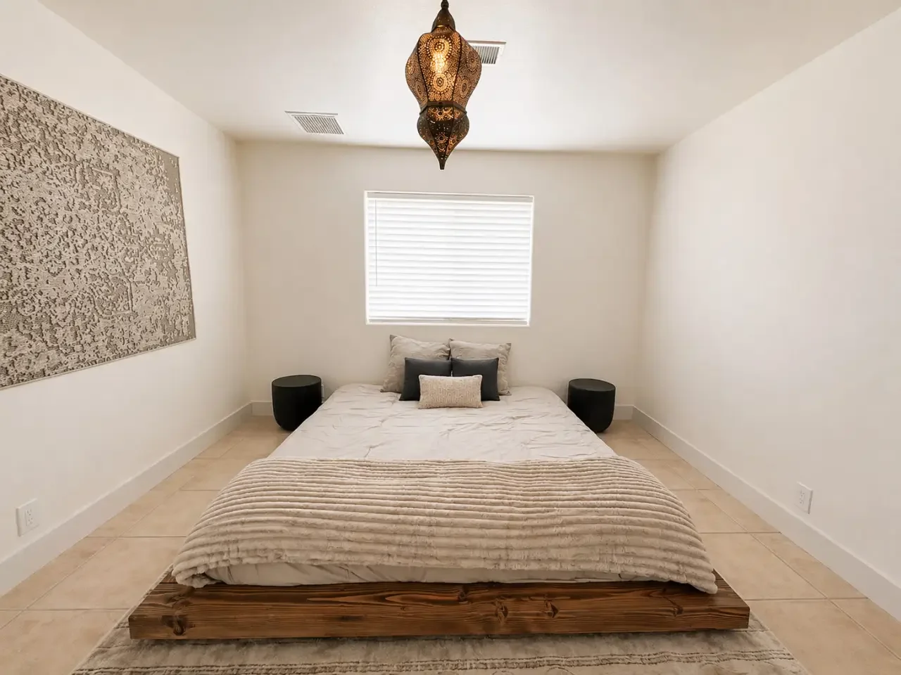 Minimalist bedroom with a low wooden platform bed, white bedding, decorative pillows, black nightstands, a textured art piece on the wall, a window with blinds, and a Moroccan-style hanging pendant light.