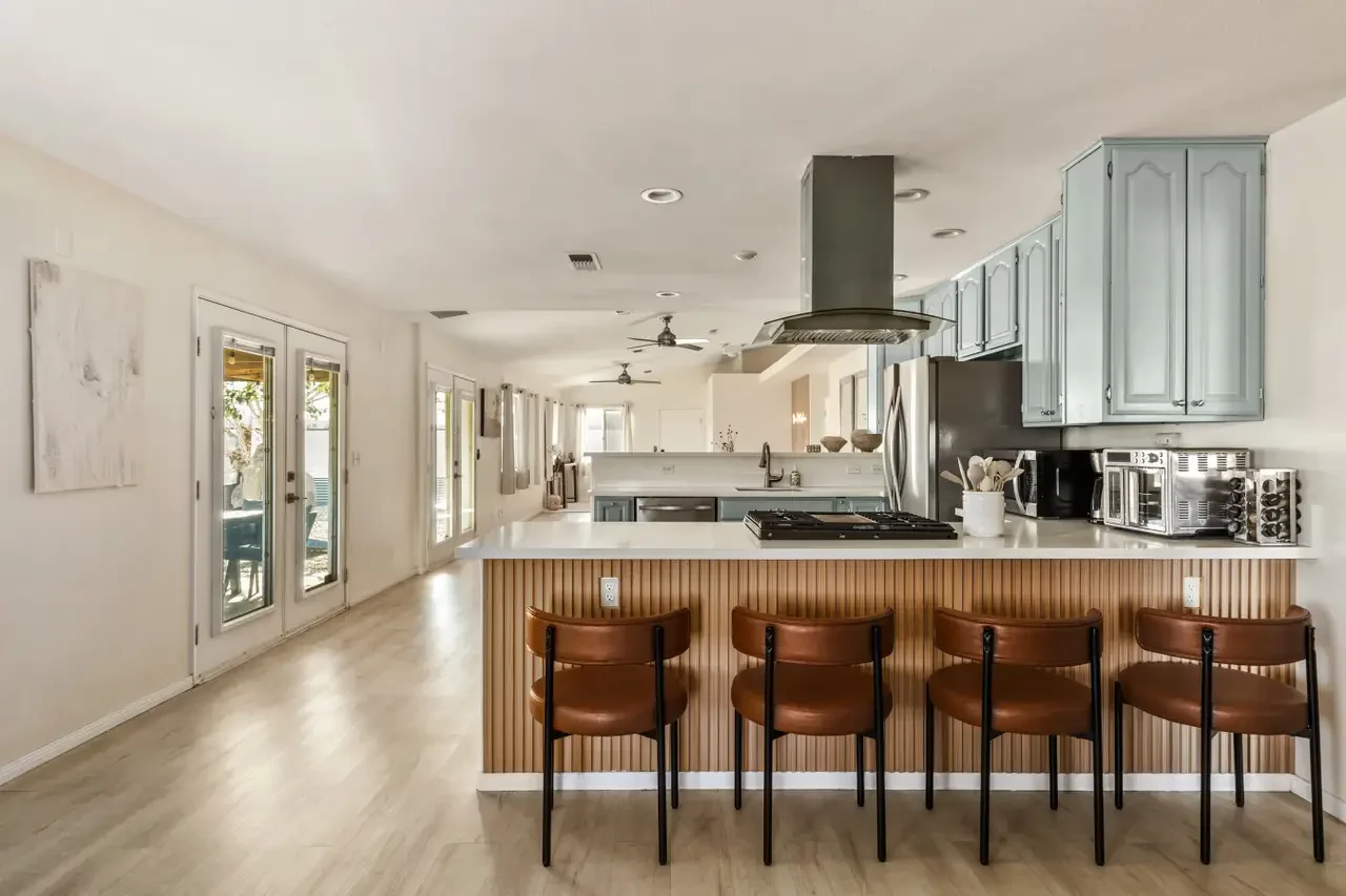 Modern kitchen with light blue cabinets, white countertops, stainless steel appliances, and four brown bar stools at a wooden kitchen island.