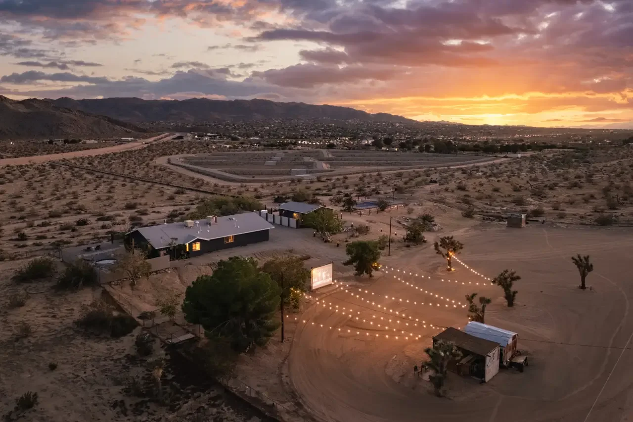 A desert landscape at sunset with a large house, string lights, and some trees.