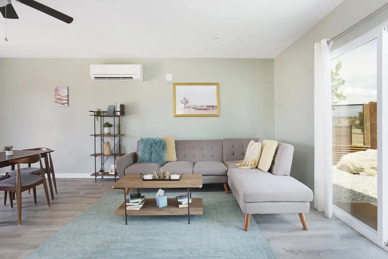 Bright living room with a sectional sofa, a coffee table with books and tissues, a bookshelf with decorative items, and a sliding glass door with white curtains revealing a yard outside.