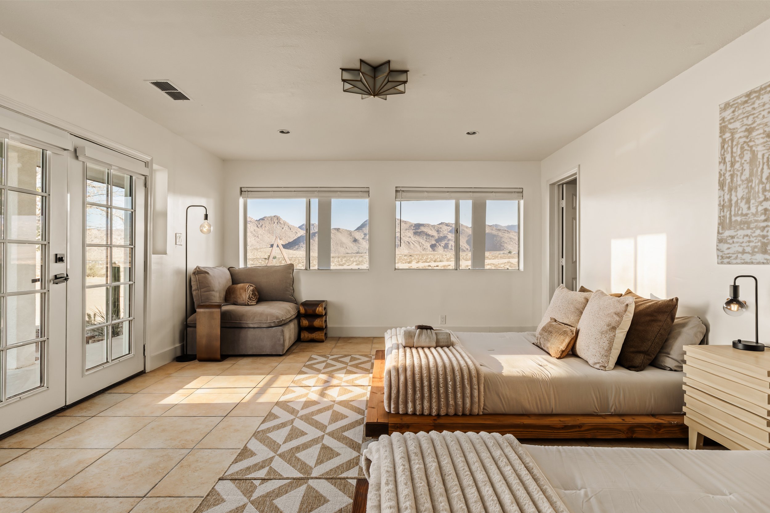 Living room with large windows showing mountainous desert landscape, beige tiled floor, beige sofa, beige armchair, patterned area rug, white walls, and modern lighting fixtures.