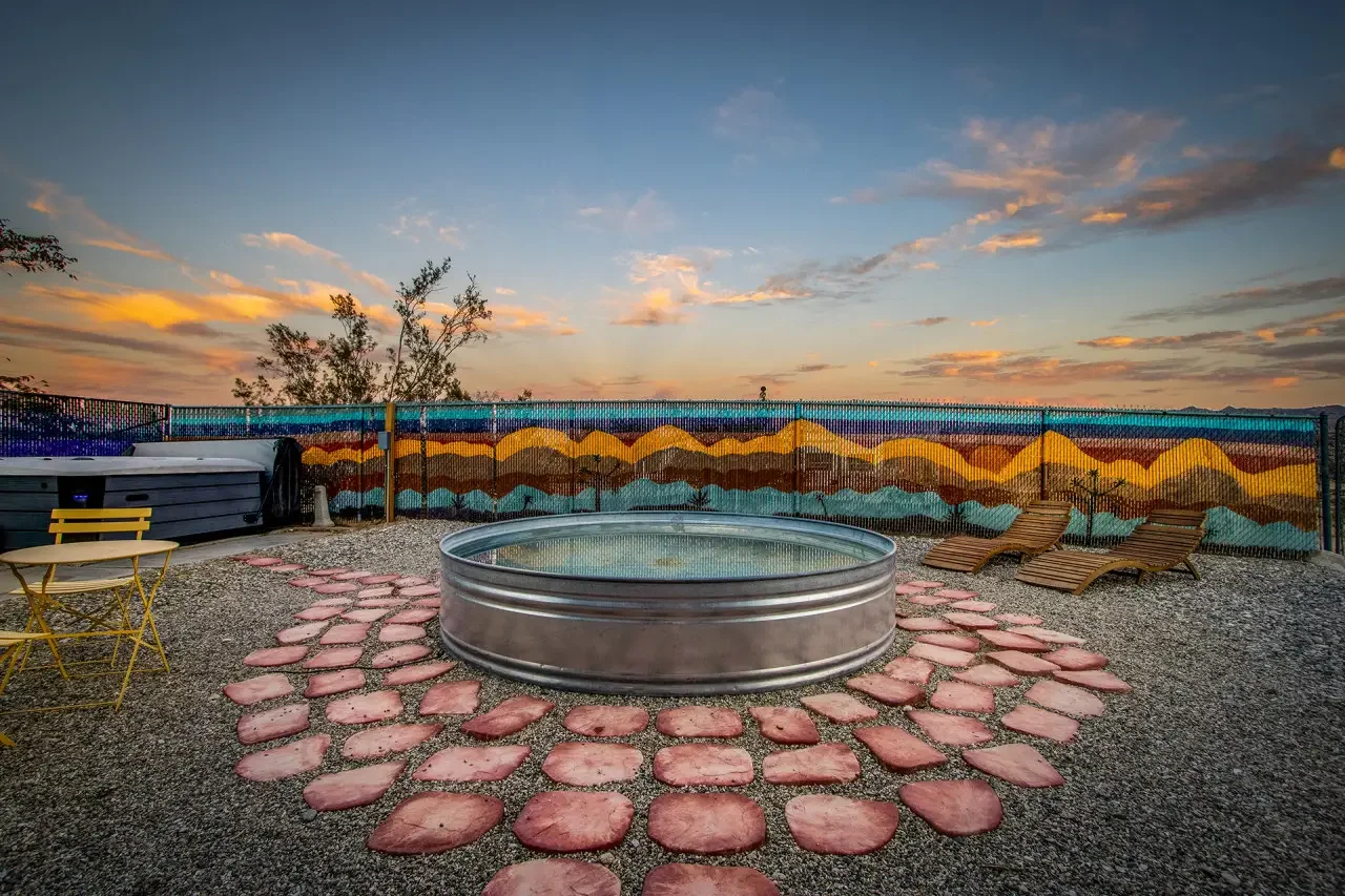 Outdoor hot tub with a circular metal exterior, surrounded by pinkish paving stones, set against a gravel ground. There are two wooden lounge chairs, a small yellow table with a matching chair, and a hot tub covering on the left. In the background, a