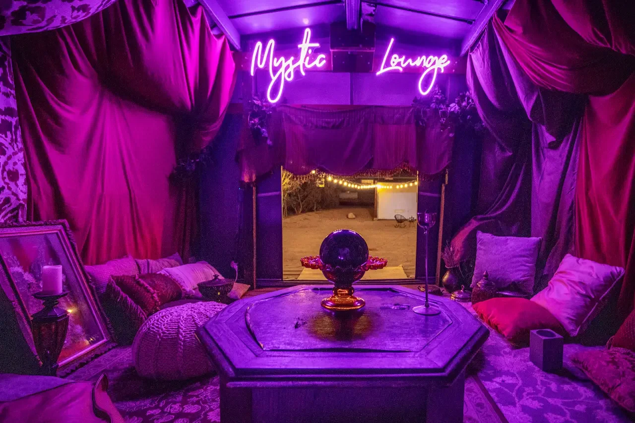 Interior of a cozy lounge with purple and pink lighting, featuring draped curtains, a wooden table, and various decorative items.