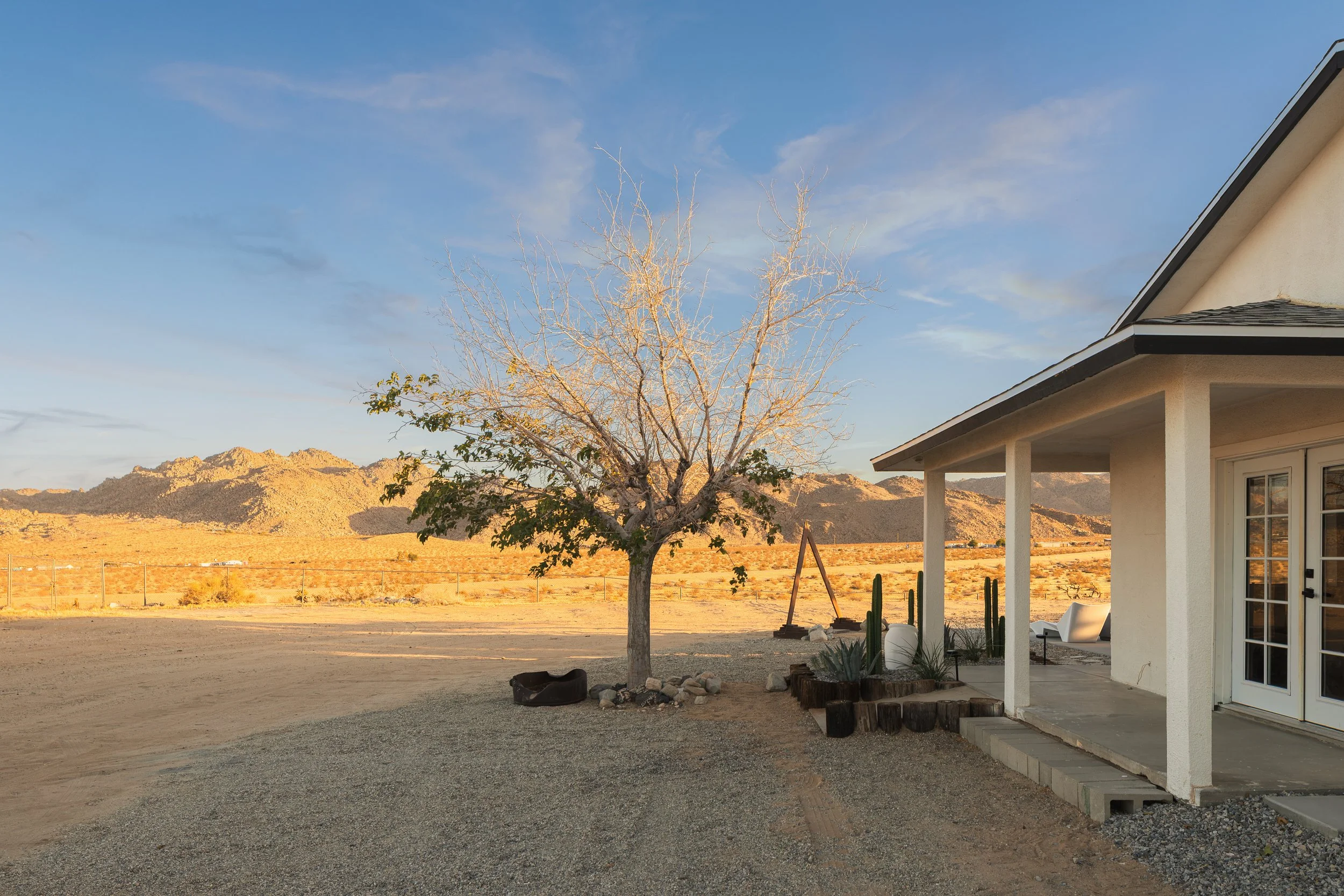A backyard scene with a house on the right, a leafless tree in the center, and mountain range in the background under a blue sky with some clouds.