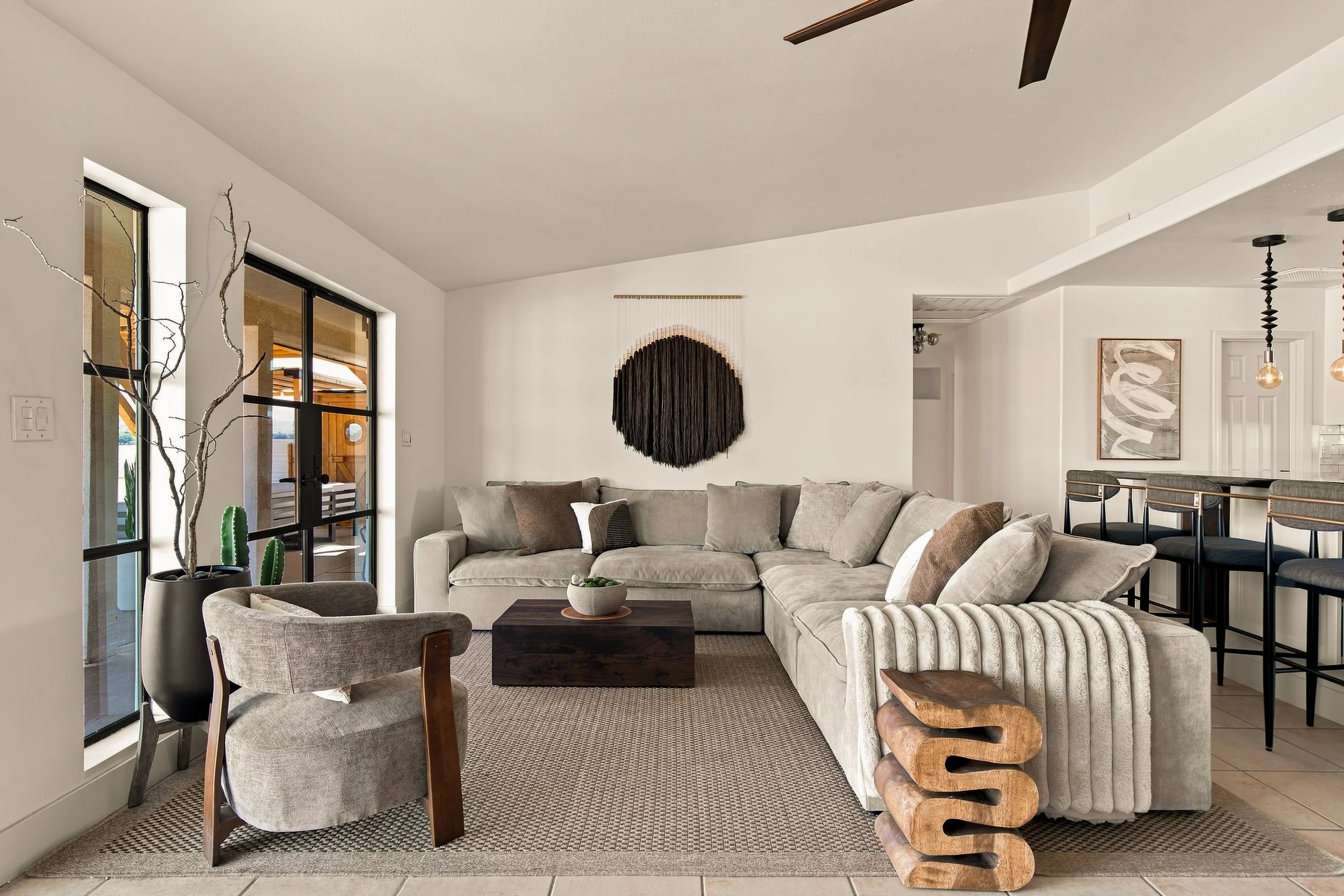 Living room with beige sectional sofa, decorative wall hanging, large windows, and bar area with stools.