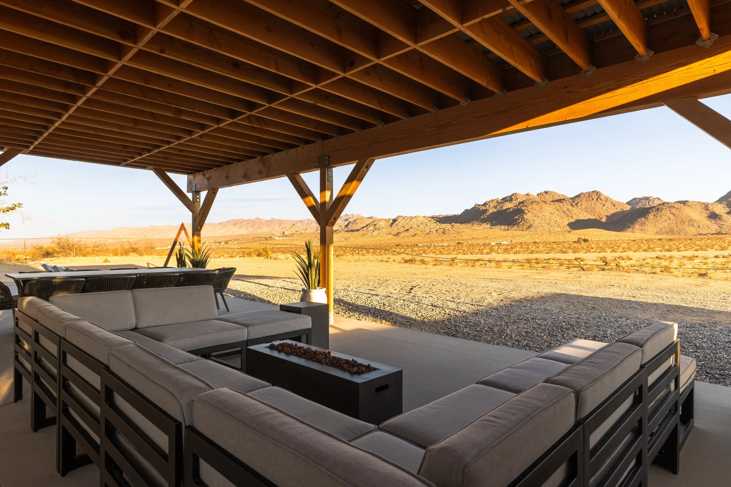 Outdoor seating area with gray sofa and coffee table under a wooden pergola, desert landscape with mountains in the background, clear blue sky.