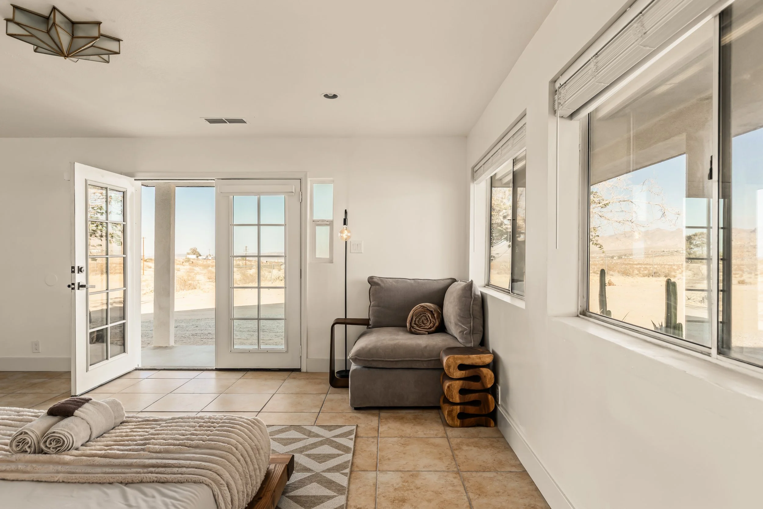 Bright living room with large windows and glass door leading outside, featuring a beige couch with rolled towels on it, decorative side table, and a beige tiled floor.
