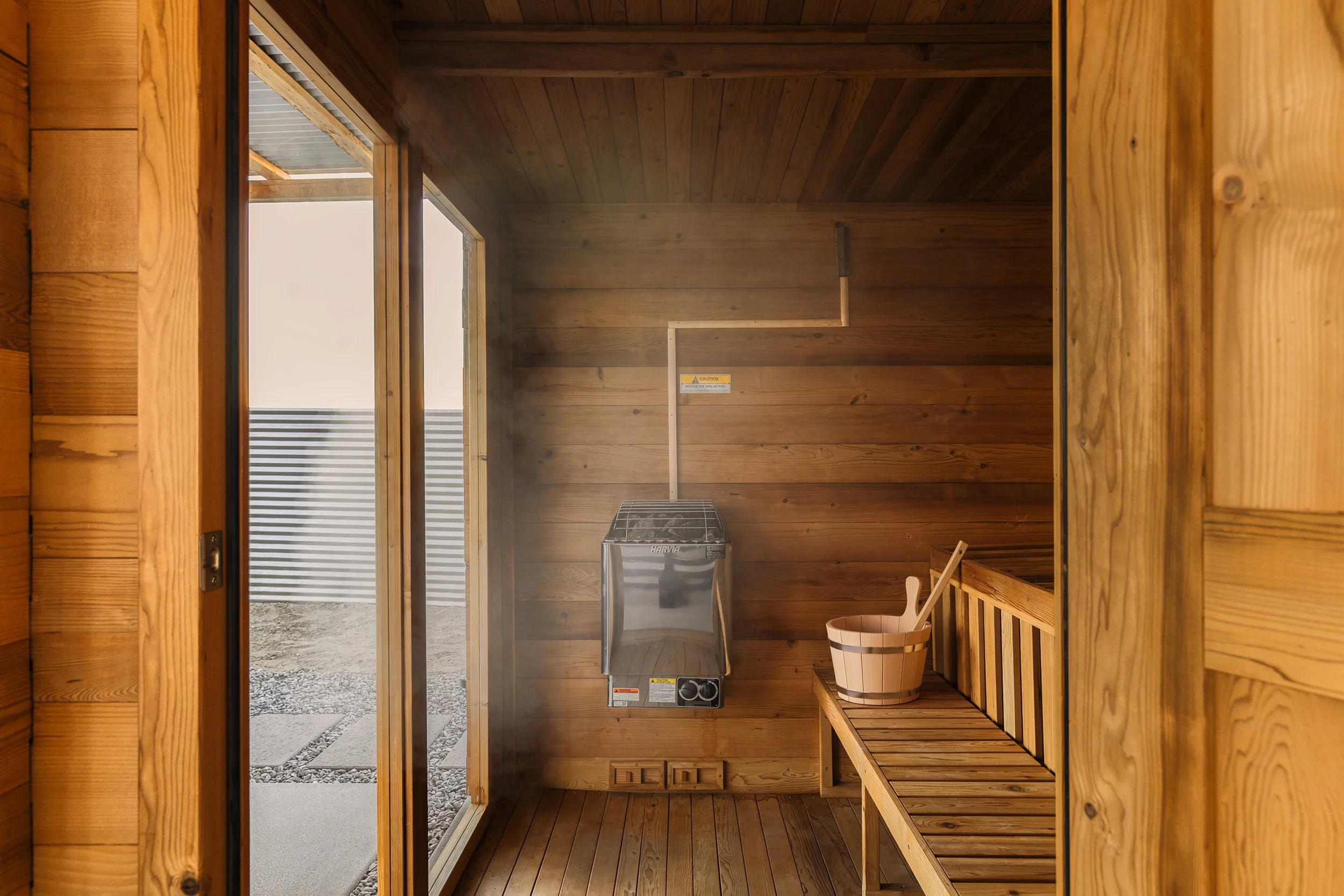 Inside a wooden sauna with benches, a wooden bucket, and a heater. The door opens to an outside area with a gravel path and metal siding.