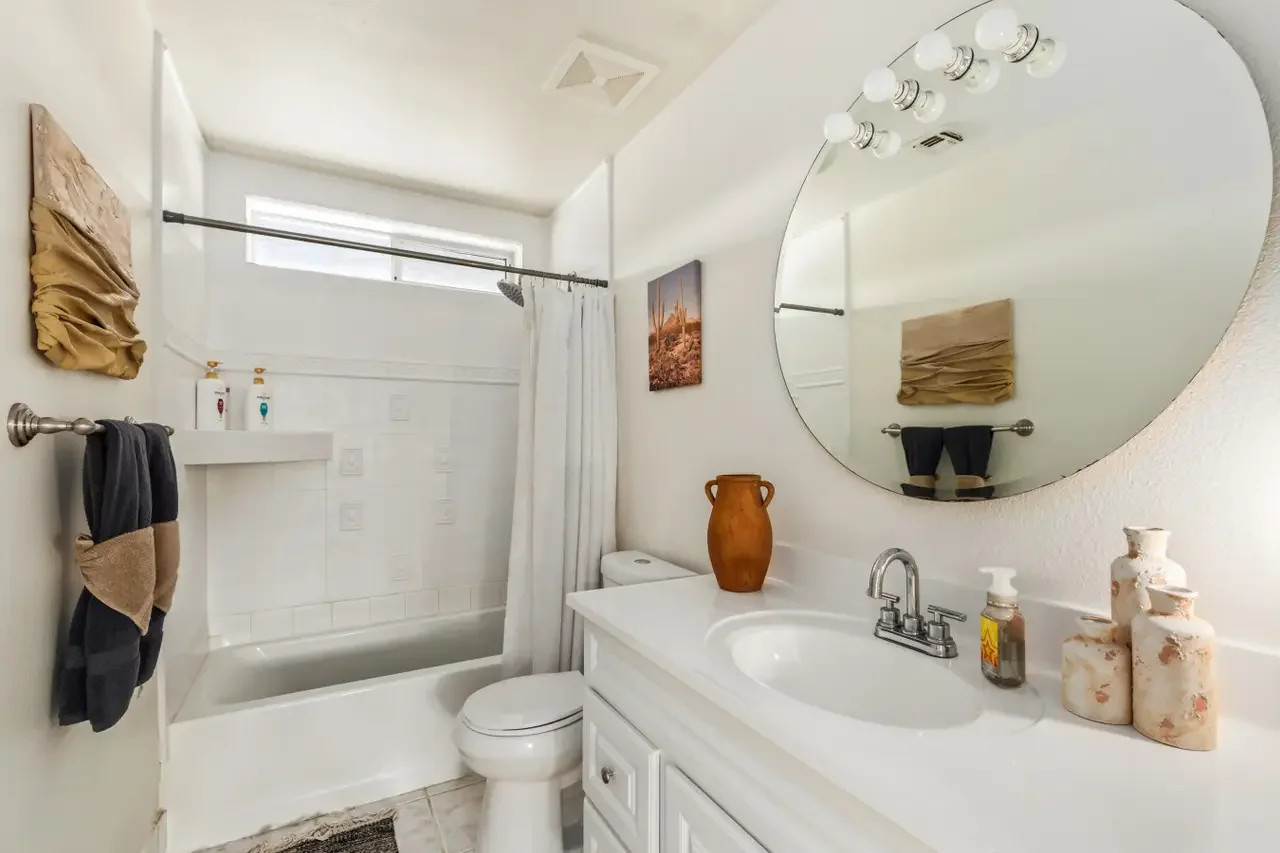 A small bathroom with a white bathtub, shower curtain, and a window above. The vanity has a round mirror, a sink with a silver faucet, and decorative bottles and vases. There is a towel rack with towels, and a piece of wall art hung on the wall.