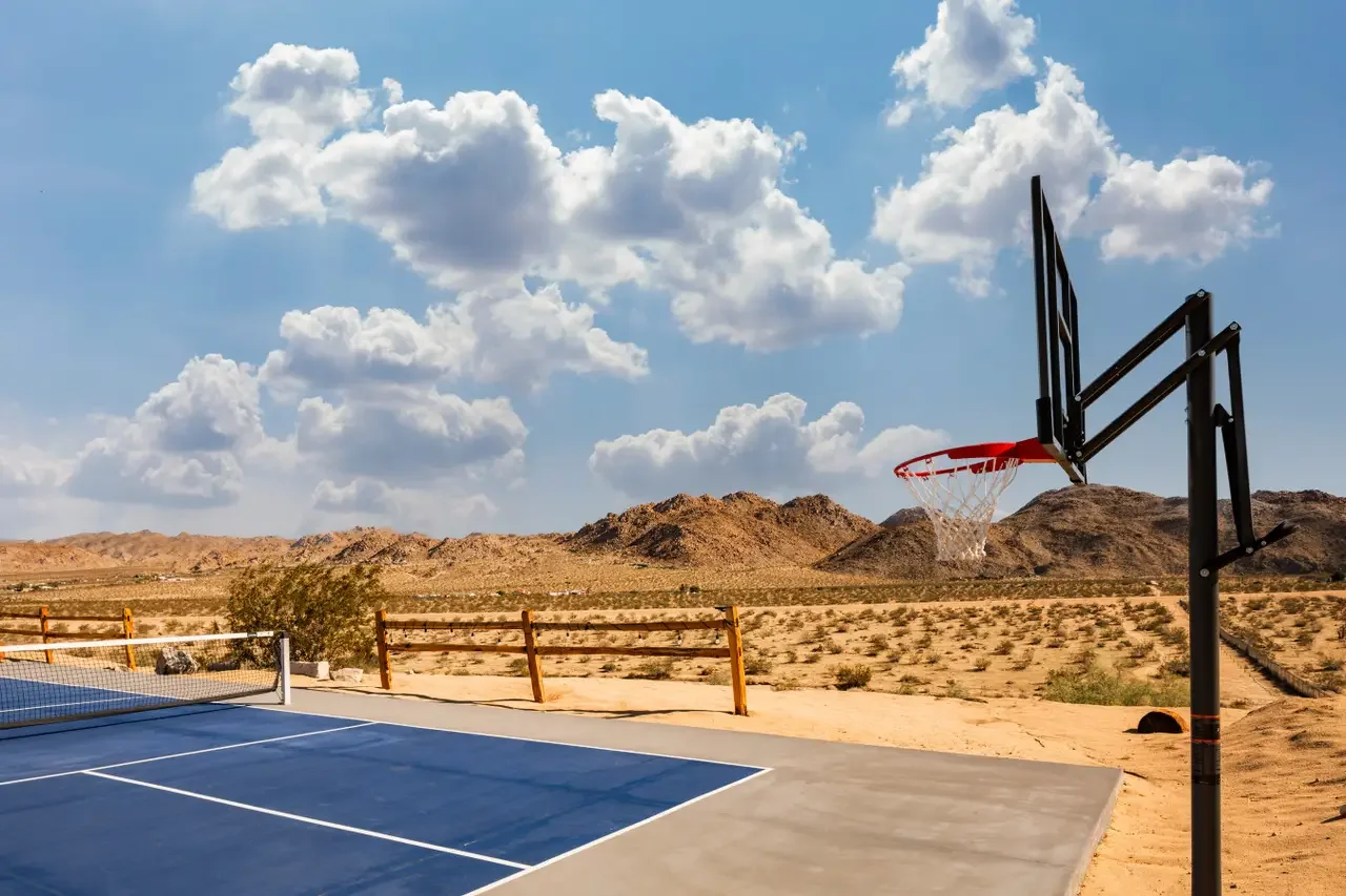 An outdoor basketball court in a desert landscape with a basketball hoop, a volleyball net, and mountains in the background, under a blue sky with scattered clouds.