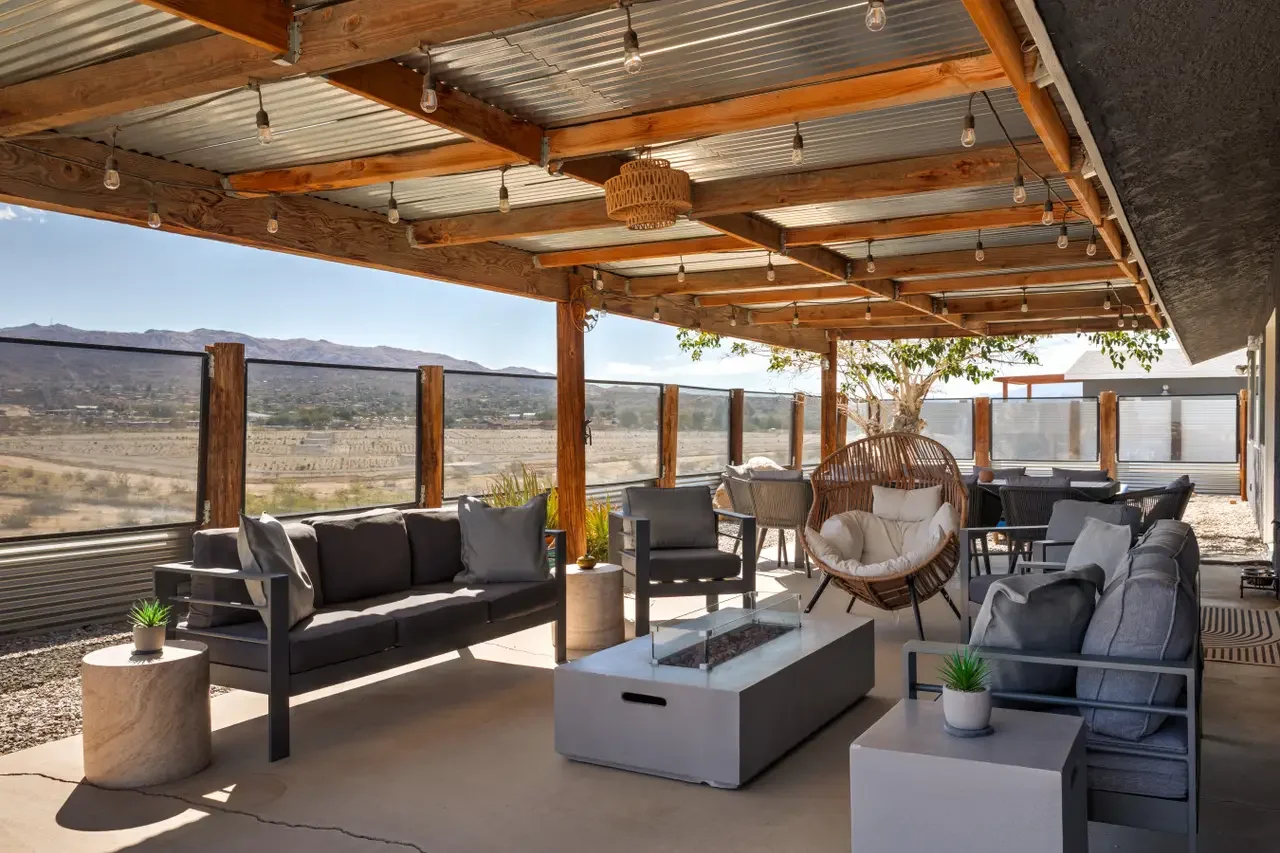 Outdoor patio with seating area including sofas, chairs, and hanging chair under a wooden pergola with string lights, overlooking a desert landscape and mountains.