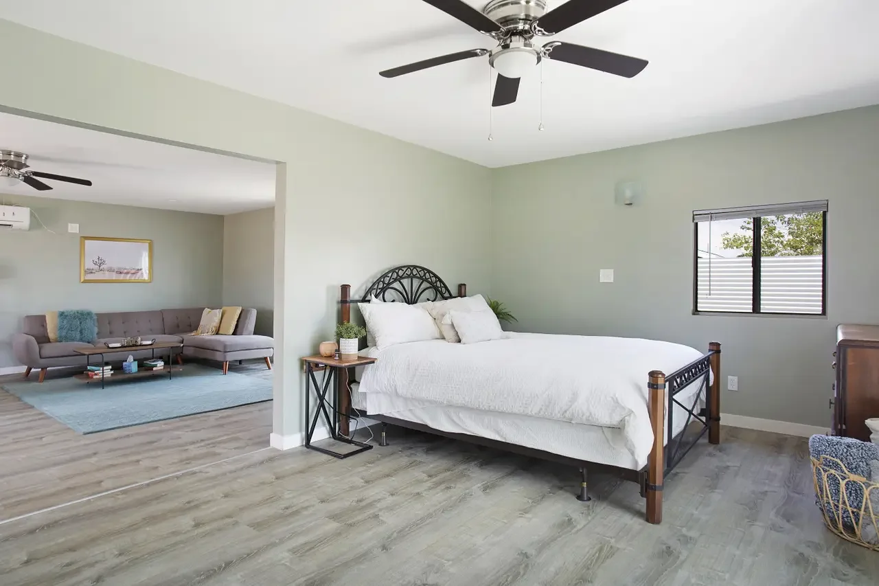 A modern bedroom with a black metal bed frame, white bedding, and a wooden nightstand with a potted plant and decorations. A window with shades shows outdoor trees. There's a transition to a living room with a gray sectional sofa, coffee table, and a
